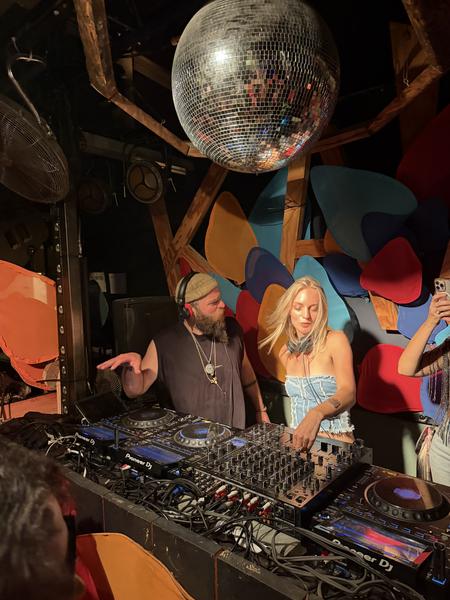 This vibrant photo captures a lively indoor music event at Zuider IJdijk, Zeeburgereiland, Amsterdam, where two DJs are actively mixing tracks on professional equipment